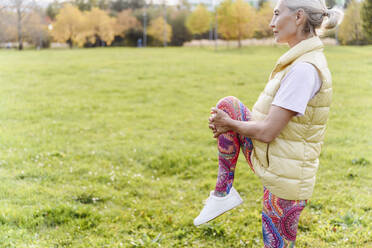 Ältere Frau, die sich auf einem Bein stehend über Gras in einem öffentlichen Park streckt - VYF00356
