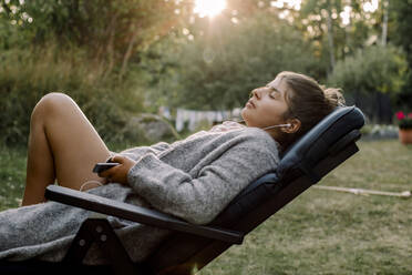 Seitenansicht eines Mädchens, das auf einem Stuhl im Garten sitzt und über sein Smartphone Musik hört - MASF20954