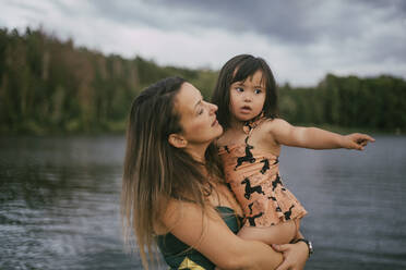 Mother carrying down syndrome daughter while standing against lake - MASF20980