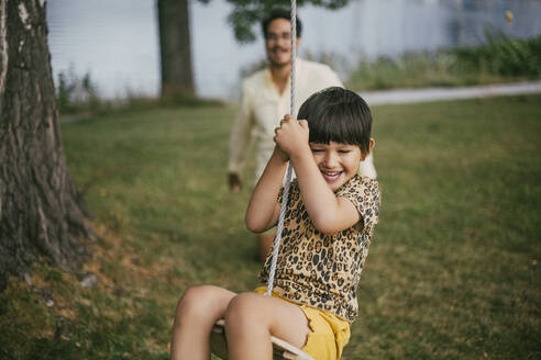 Lächelnde Tochter sitzt auf einer Seilschaukel, während der Vater im Hintergrund im Garten steht - MASF20984
