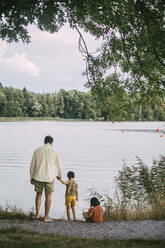 Rear view of father holding hands with daughter standing against lake - MASF20987
