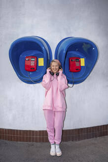 Lächelnde junge Frau mit rosa Haaren und rosa Kapuzenshirt vor einer Telefonzelle stehend - VPIF03283