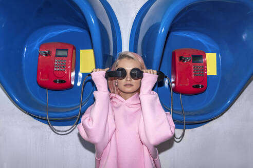 Young woman with pink hair wearing pink hooded shirt standing at telephone booths, holding receivers in front of eyes - VPIF03286