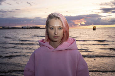 Porträt einer jungen Frau mit rosa Haaren und rosa Kapuzenshirt mit Meer bei Sonnenuntergang im Hintergrund - VPIF03297