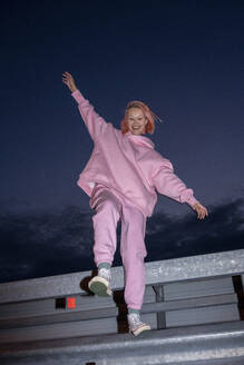 Junge Frau mit rosa Haaren und rosa Trainingsanzug, die auf einem Bein auf einer Straßenschranke in der Nacht steht - VPIF03312