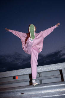 Junge Frau in rosafarbenem Trainingsanzug steht nachts auf einem Bein auf einer Straßenschranke - VPIF03313