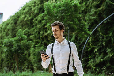 Businessman wearing white shirt and black suspenders looking at smart phone outdoors - XLGF00860