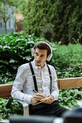 Young businessman with headphones sitting on bench outdoors and writing in notepad - XLGF00863