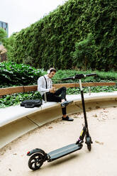 Young businessman with headphones sitting on bench outdoors and writing in notepad - XLGF00864