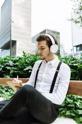 Junger Geschäftsmann mit Kopfhörern sitzt auf einer Bank im Freien und schreibt in einen Notizblock - XLGF00865