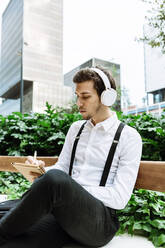 Young businessman with headphones sitting on bench outdoors and writing in notepad - XLGF00865