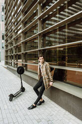 Stylish man in check jacket with push scooter sitting at modern building in city - XLGF00871