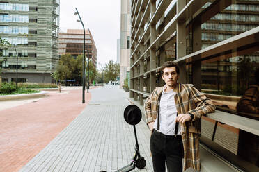 Porträt eines stilvollen Mannes in karierter Jacke mit einem Motorroller vor einem modernen Gebäude in der Stadt - XLGF00872
