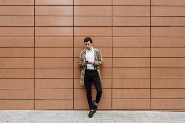 Stilvoller Mann in karierter Jacke, der an der Wand lehnt und sein Smartphone benutzt - XLGF00875