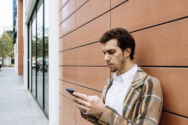 Stylischer junger Mann mit Smartphone vor einem Gebäude stehend - XLGF00879