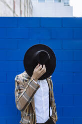 Stilvoller Mann mit karierter Jacke, der einen schwarzen Hut vor das Gesicht hält, vor einer blauen Wand - XLGF00890