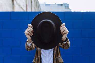 Stilvoller Mann mit karierter Jacke, der einen schwarzen Hut vor das Gesicht hält, vor einer blauen Wand - XLGF00891