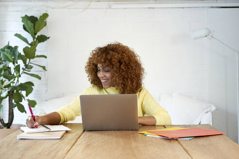 Happy businesswoman with laptop writing in notepad while working from home - VEGF03305