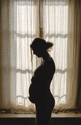 Pregnant woman touching belly while standing by window at home - MRAF00580