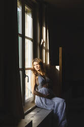 Schwangere Frau träumt beim Blick durch das Fenster im Sonnenlicht zu Hause - MRAF00582