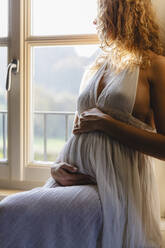 Pregnant woman touching belly while leaning on window at home - MRAF00585