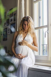 Young pregnant woman looking at abdomen while sitting on window seat at home - MRAF00586