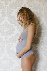 Schwangere Frau, die sich zu Hause an der Wand stehend um ihren Unterleib kümmert - MRAF00588