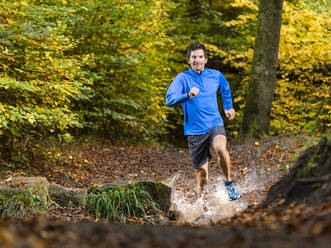 Lächelnde männliche Sportlerin beim Trailrunning, während sie Wasser aus dem Bach am Kappelberg spritzt, Deutschland - STSF02726