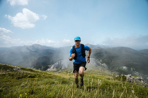 Mature sportsman trail running in the mountains on meadow against blue sky - JMPF00712