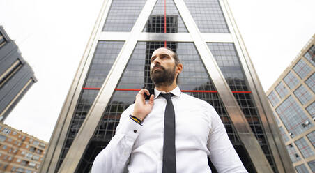 Porträt eines bärtigen Geschäftsmannes, der vor einem modernen Wolkenkratzer steht - JCCMF00188