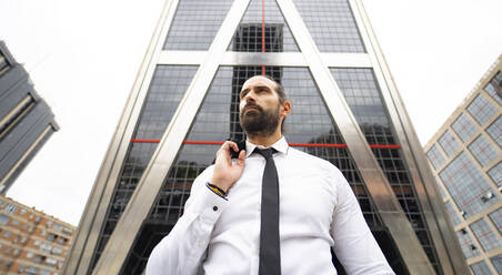 Portrait of bearded businessman standing against modern skyscraper - JCCMF00188