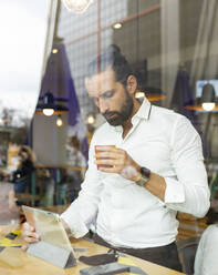 Candid portrait of bearded businessman drinking coffee in front of digital tablet inside cafe - JCCMF00190