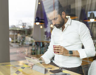 Unverfälschtes Porträt eines bärtigen Geschäftsmannes, der vor einem digitalen Tablet in einem Café Kaffee trinkt - JCCMF00191