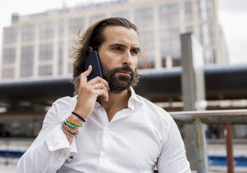 Candid portrait of bearded businessman sitting outdoors and talking on smart phone - JCCMF00197