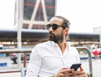 Portrait of bearded businessman wearing sunglasses sitting outdoors with smart phone in hands - JCCMF00198
