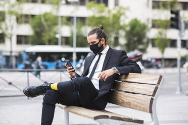 Geschäftsmann mit Schutzmaske auf einer Bank sitzend mit Smartphone in der Hand - JCCMF00205