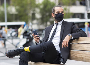 Porträt eines Geschäftsmannes mit Gesichtsschutzmaske, der auf einer Bank sitzt und ein Smartphone in der Hand hält - JCCMF00207