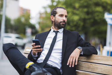 Porträt eines bärtigen Geschäftsmannes, der auf einer Bank sitzt und ein Smartphone in der Hand hält - JCCMF00209