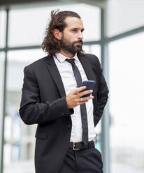 Porträt eines bärtigen Geschäftsmannes, der mit einem Smartphone in der Hand in einem Haus steht - JCCMF00212