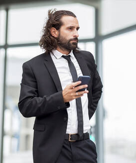 Portrait of bearded businessman standing indoors with smart phone in hand - JCCMF00212