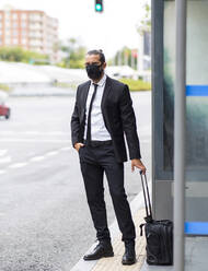 Businessman wearing protective face mask waiting at bus stop with suitcase - JCCMF00215