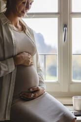 Pregnant woman with hands on stomach sitting by window at home - MRAF00592