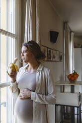 Junge schwangere Frau, die eine Banane isst, während sie durch ein Fenster zu Hause schaut - MRAF00593