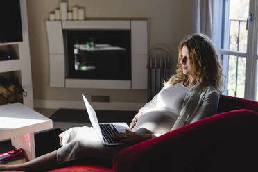 Pregnant freelancer using laptop while sitting on sofa in living room - MRAF00595