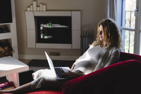 Schwangere Freiberuflerin, die einen Laptop benutzt, während sie auf dem Sofa im Wohnzimmer sitzt - MRAF00595