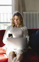Geschäftsfrau mit Laptop, die ihr Smartphone benutzt, während sie auf dem Sofa im Wohnzimmer sitzt - MRAF00597