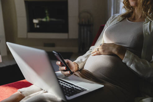 Schwangere Geschäftsfrau mit Mobiltelefon und Laptop auf dem Sofa zu Hause - MRAF00598