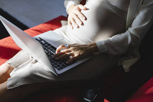 Schwangere Freiberuflerin tippt zu Hause auf dem Laptop - MRAF00599