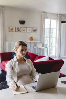 Freiberuflerin, die in ein Buch schreibt, während sie einen Laptop am Schreibtisch im Wohnzimmer benutzt - MRAF00600
