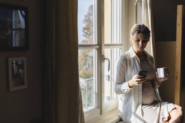 Schwangere Frau mit Kaffeetasse Textnachrichten auf Handy gegen Fenster - MRAF00605